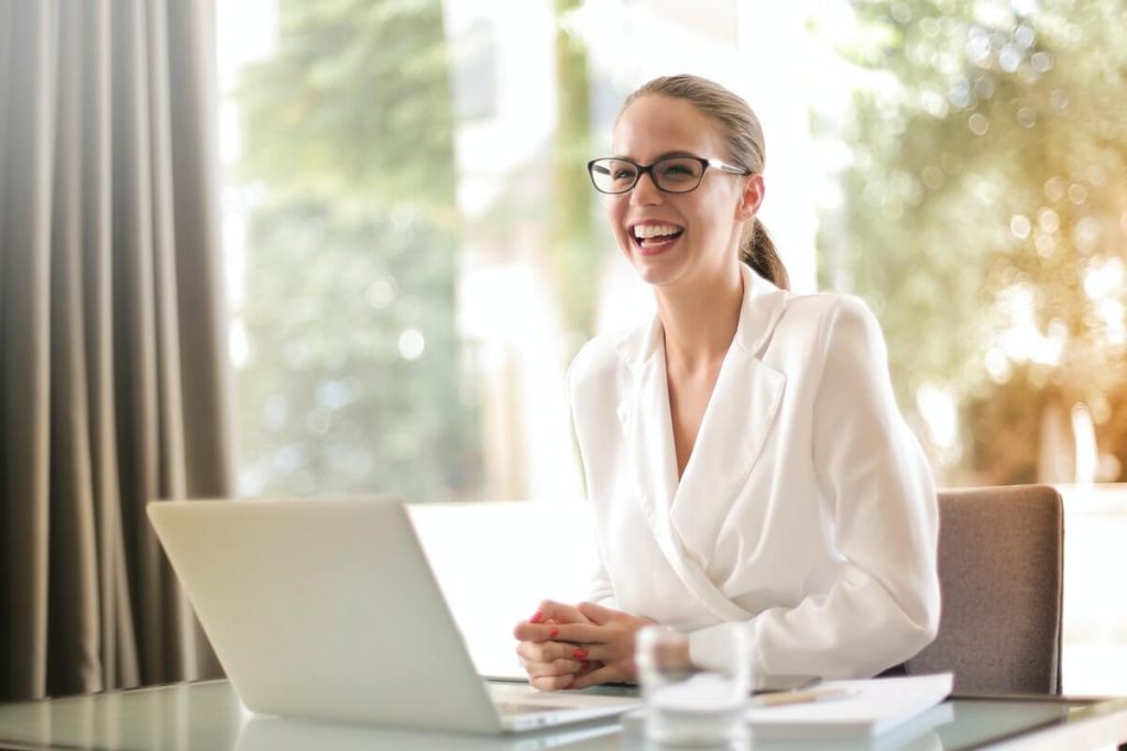 Eine Frau in einem weißem Blazer gekleidet lächelt freundlich neben ihrem Notebook vor einem hellen Hintergrund.
